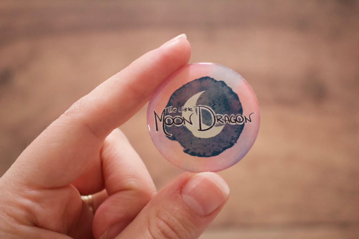 A hand holding a round sticker featuring a watercolor-style design with the text 'The Little Moon Dragon'.