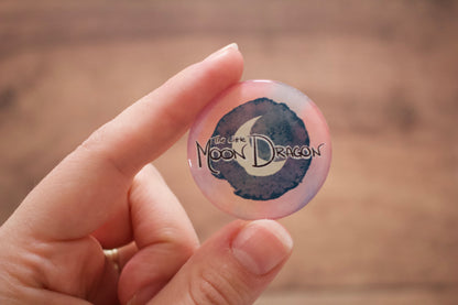 A hand holding a round sticker featuring a watercolor-style design with the text 'The Little Moon Dragon'.