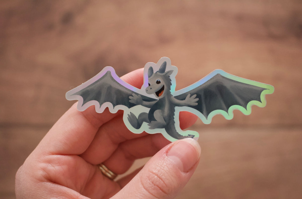 A hand holding a die-cut sticker of a cute gray cartoon dragon with spread wings and a holographic iridescent border, set against a warm, blurred wooden background.