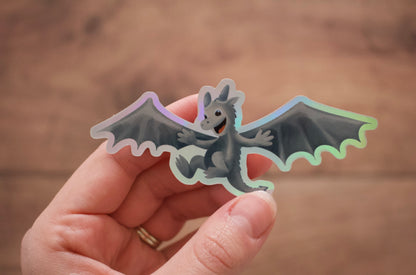 A hand holding a die-cut sticker of a cute gray cartoon dragon with spread wings and a holographic iridescent border, set against a warm, blurred wooden background.