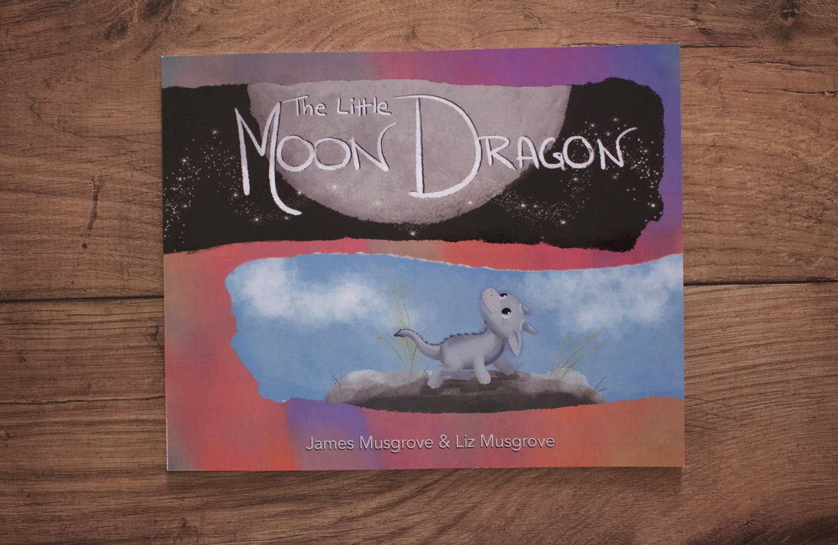 The Little Moon Dragon children's book cover displayed flat on a rustic wooden surface. The cover features a whimsical illustration of a small gray dragon under a large moon, with a black starry sky on top and a blue sky with clouds on the bottom.