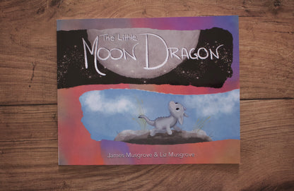 The Little Moon Dragon children's book cover displayed flat on a rustic wooden surface. The cover features a whimsical illustration of a small gray dragon under a large moon, with a black starry sky on top and a blue sky with clouds on the bottom.